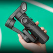 Hand holding black smartphone gimbal stabilizer against teal background